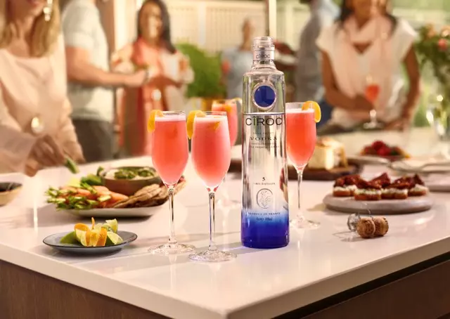 A bottle of Ciroc vodka and three pink cocktails with lemon garnishes on a kitchen counter surrounded by plates of food, with people in the background.