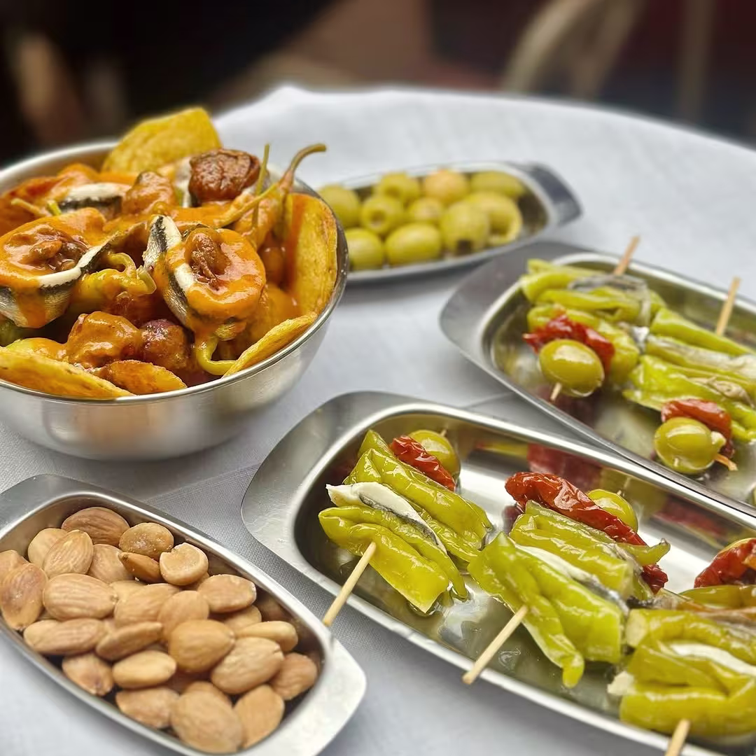 La imagen muestra una selección de tapas que incluye patatas con salsa, aceitunas, almendras y brochetas de pimientos con tomates secos, todo dispuesto en platos metálicos sobre una mesa blanca.