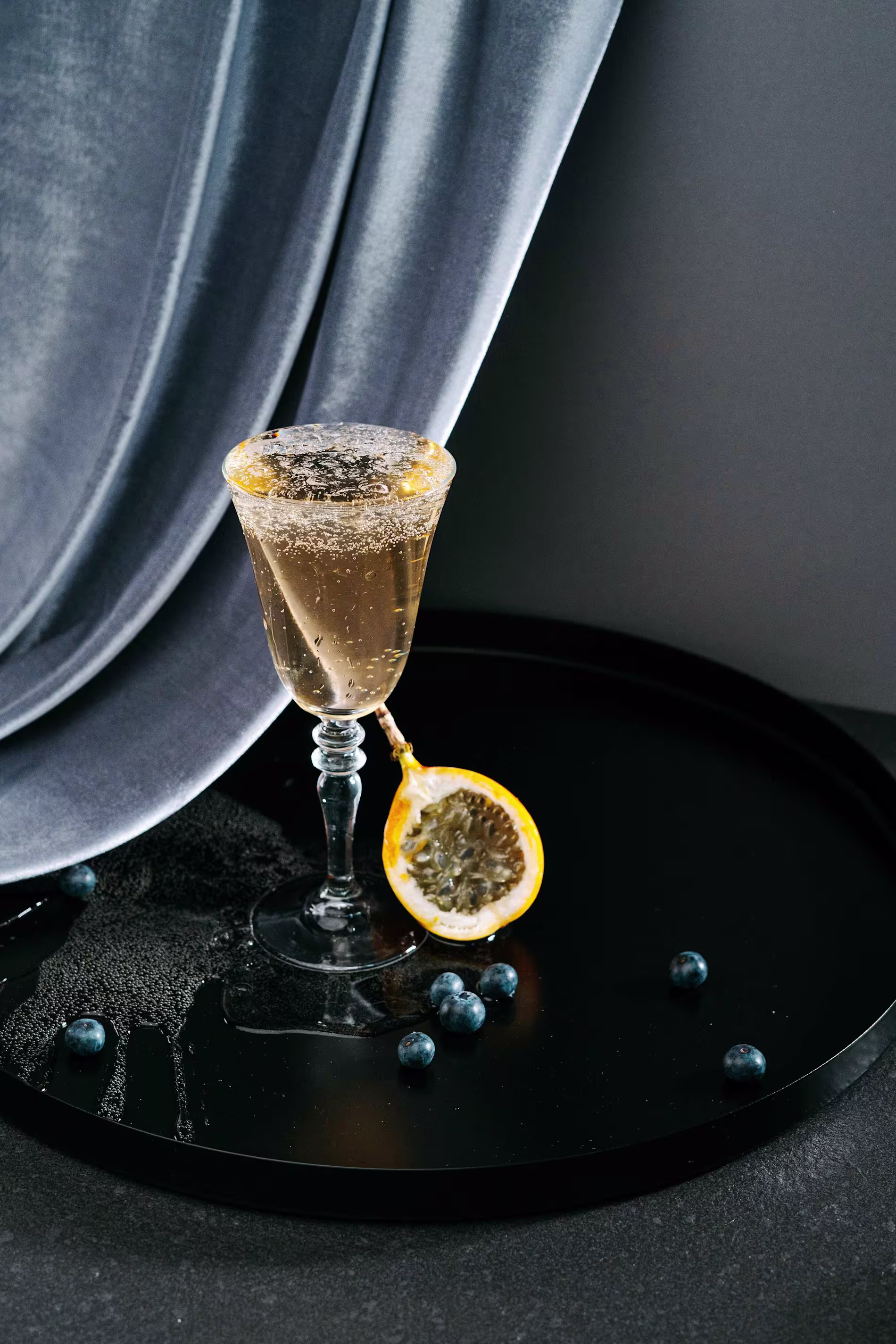 A glass of sparkling drink with a sugared rim sits on a black tray, next to a halved passion fruit and scattered blueberries, with a gray fabric backdrop.