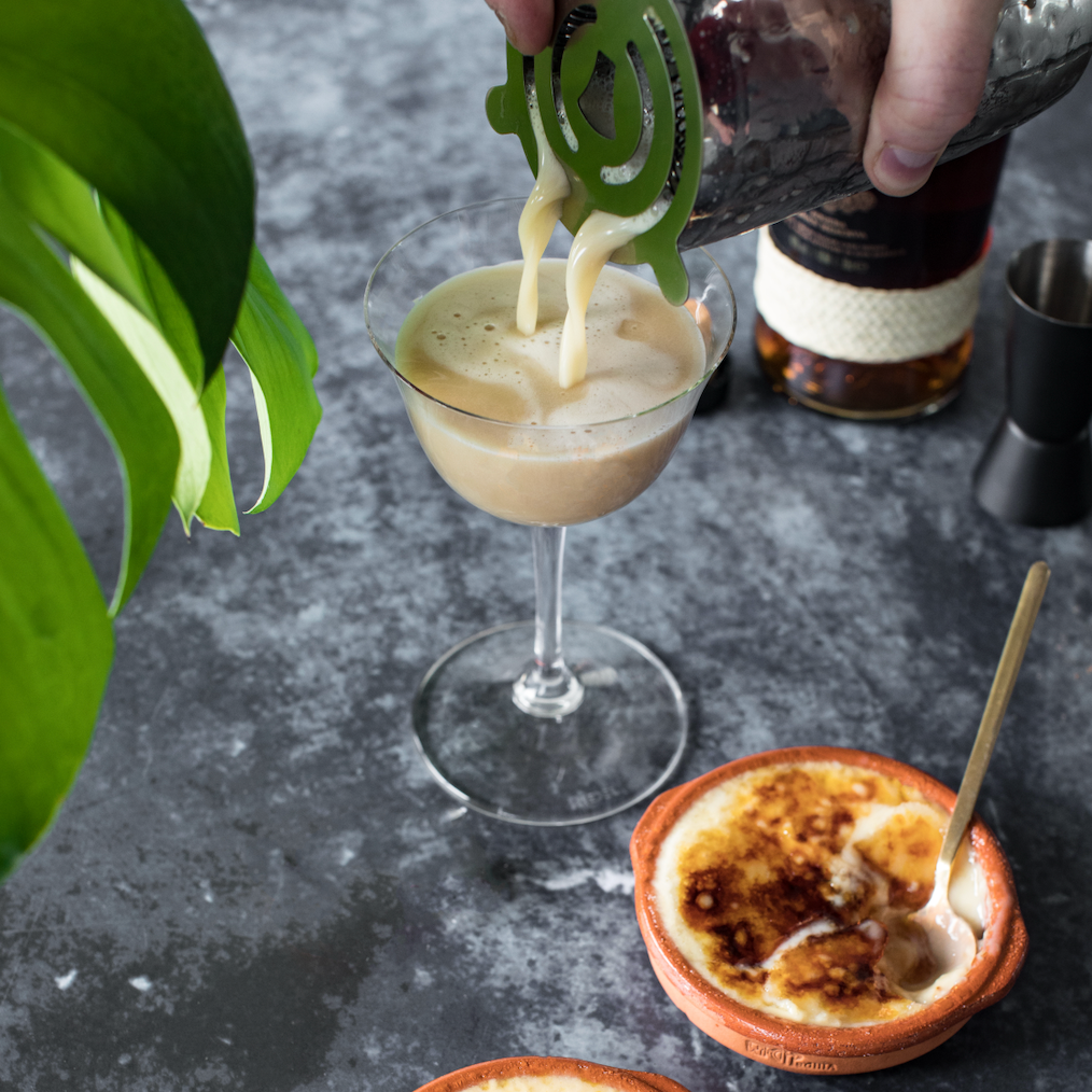 A bartender pours a rum cocktail from a silver cocktail shaker into a coupe glass.