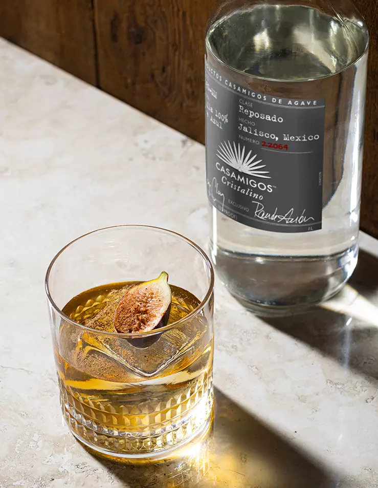 A glass of amber liquor with a fig garnish sits on a marble surface next to a bottle of Casamigos Cristalino tequila.
