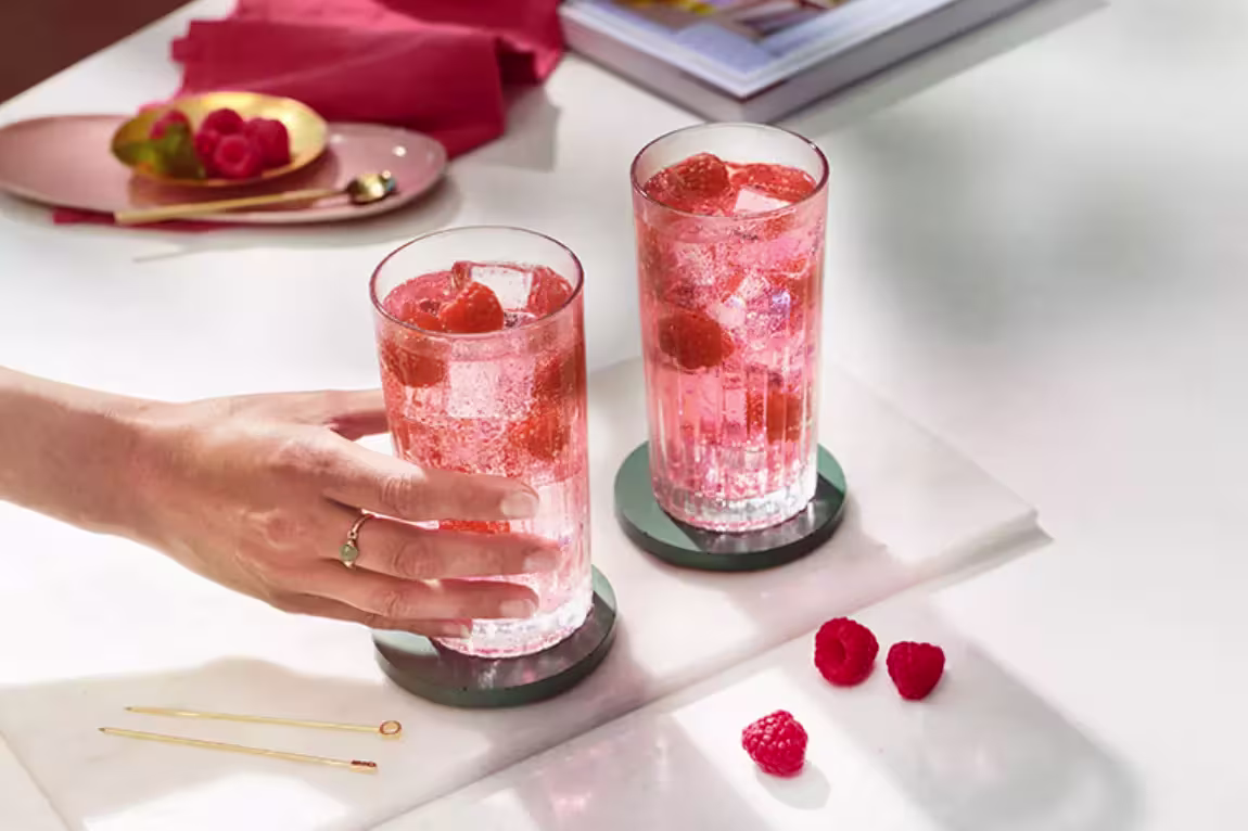 Two tall glasses of pink fizzy drink with ice and raspberries on coasters; a hand takes one glass. Raspberries and cocktail picks are on the table nearby.