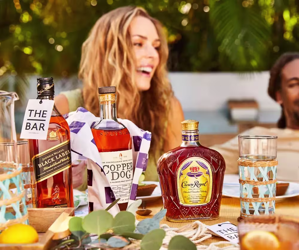 Three bottles of liquor and glasses are arranged on an outdoor table, with two people smiling in the background.