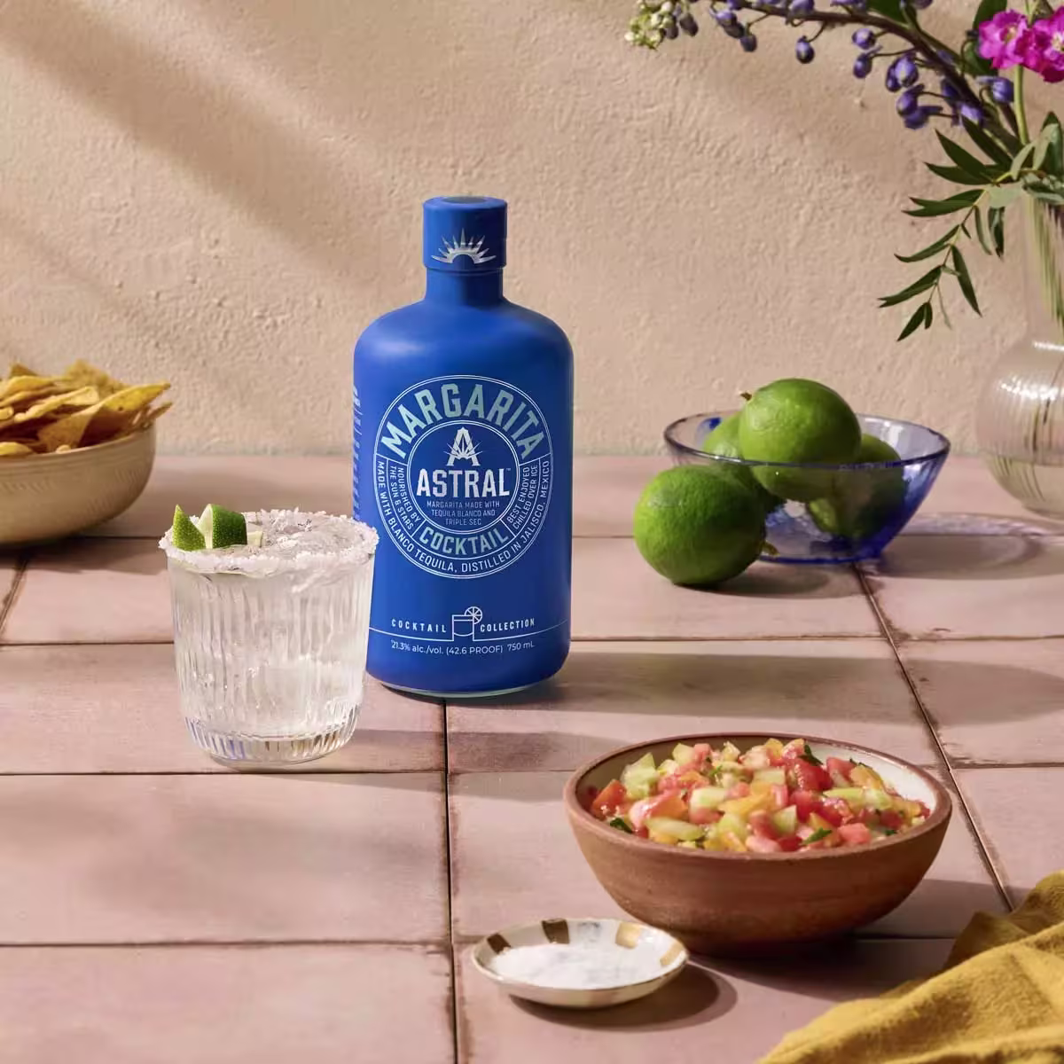 A bottle of Astral Margarita, a salt-rimmed margarita glass with lime, a bowl of salsa, limes in a glass bowl, chips, and a small dish of salt on a tiled table.