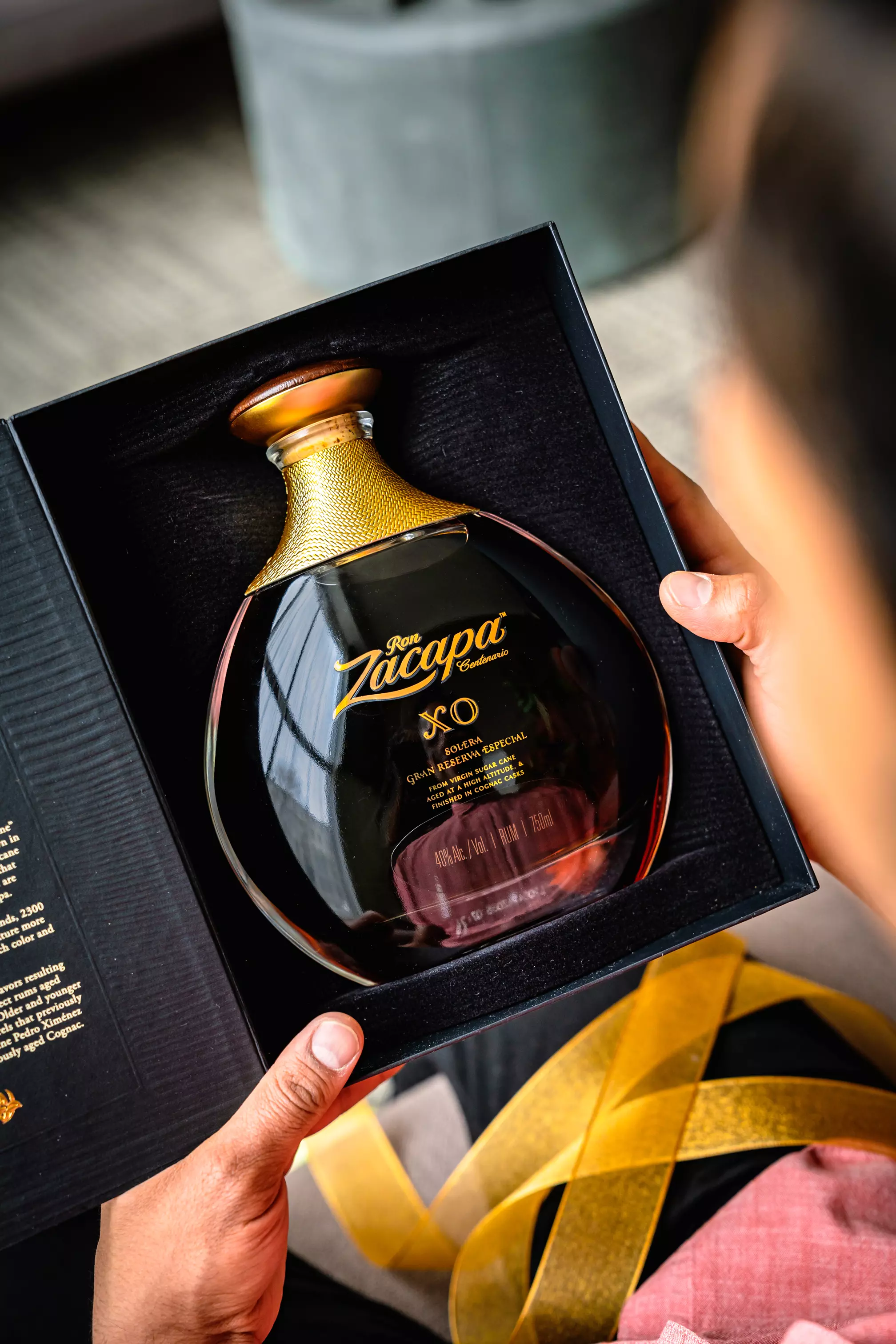 A person holds an open box containing a bottle of Ron Zacapa XO rum with gold lettering and a gold cap.