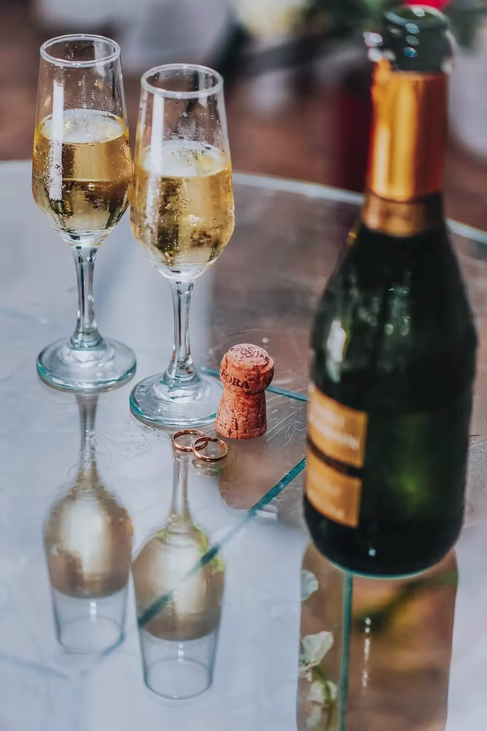 Two champagne glasses and a bottle sit on a glass table with a cork and two wedding rings nearby.