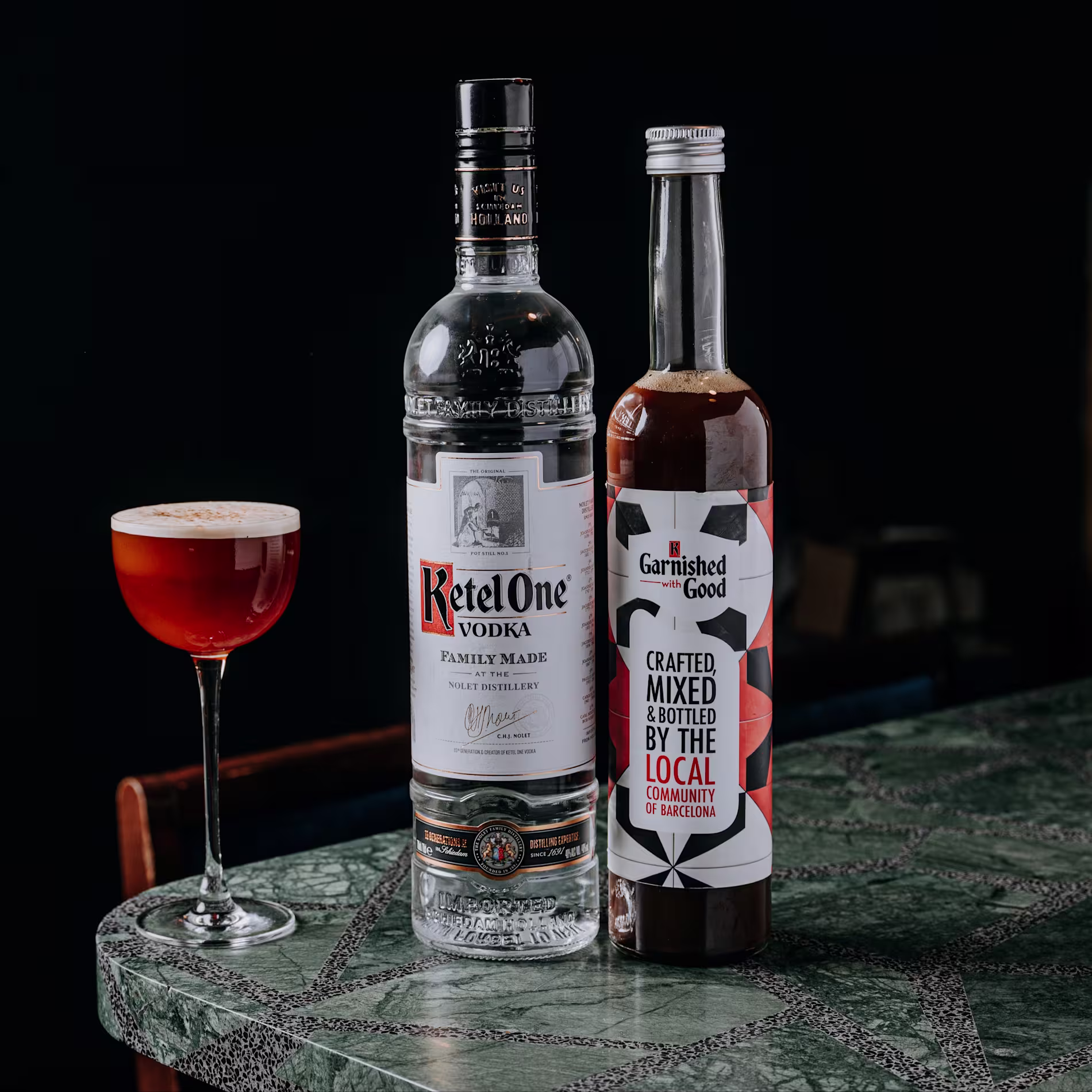 Cóctel servido en copa junto a una botella de Ketel One Vodka y otra de mezcla artesanal. Todo sobre una barra verde con fondo oscuro y ambiente elegante.