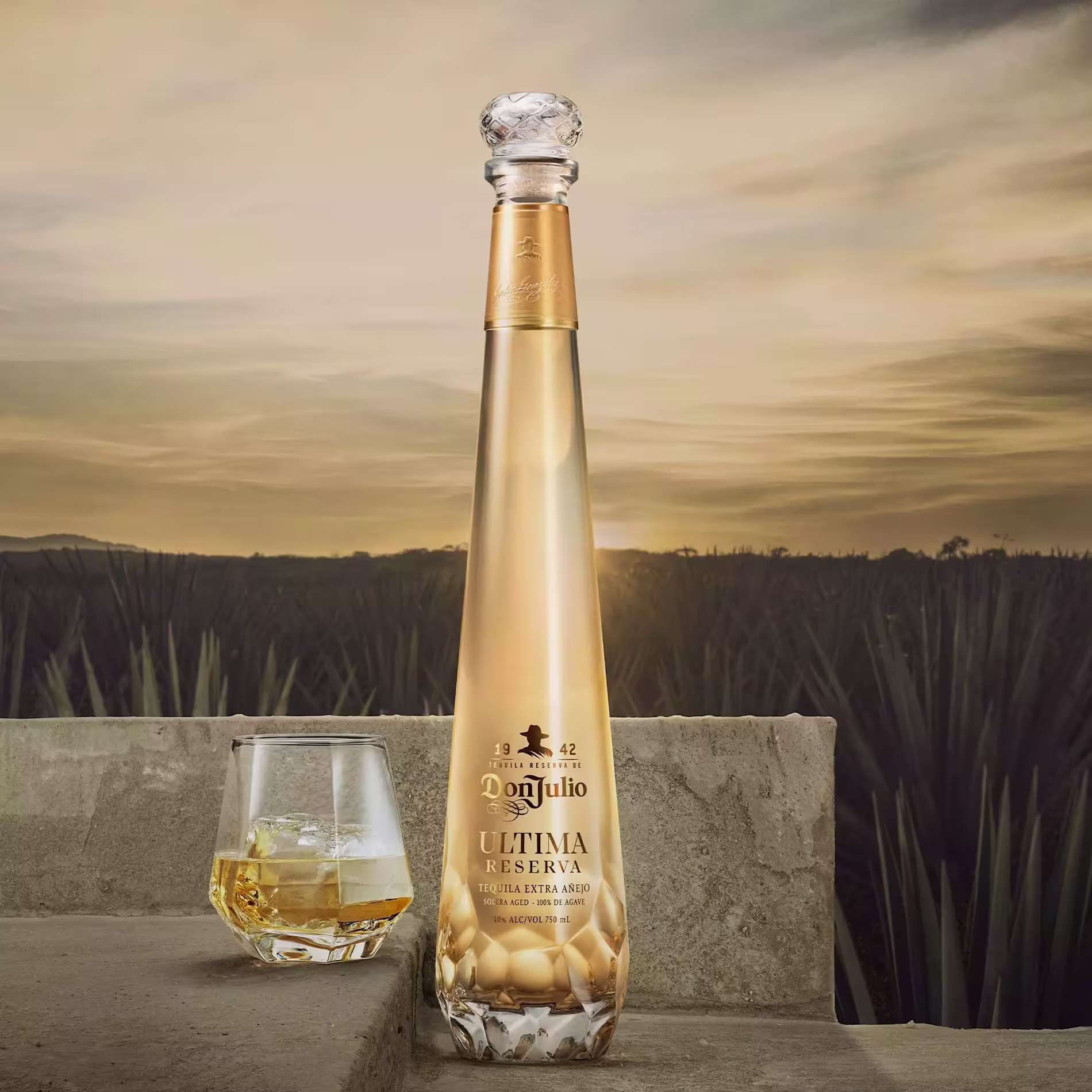 A tall bottle of Don Julio Ultima Reserva tequila stands next to a glass of tequila with ice, set against an agave field at sunset.
