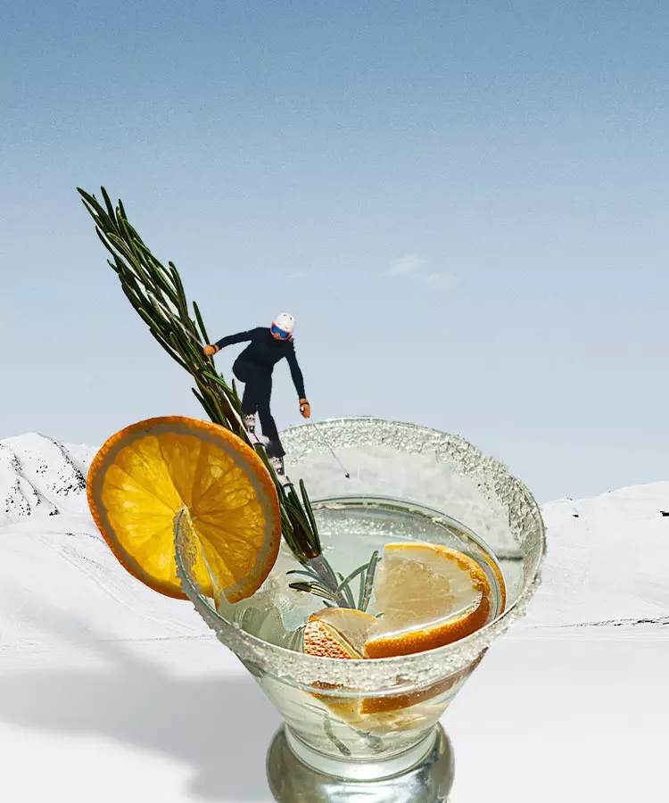 A skier goes downhill in the snowy mountains, with a cocktail glass containing citrus and rosemary in the foreground.