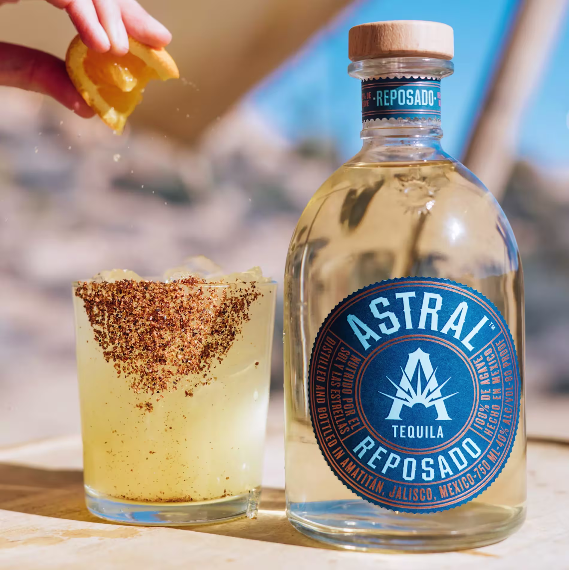 A bottle of Astral Reposado Tequila next to a cocktail in a glass rimmed with chili powder, with a hand squeezing an orange peel over the drink.
