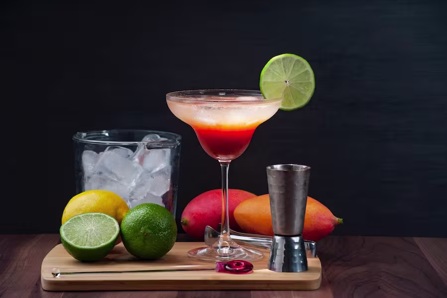 A cocktail with a lime slice garnish sits on a board with a jigger, bar spoon, lime, lemon, mangoes, and a glass container of ice against a dark background.