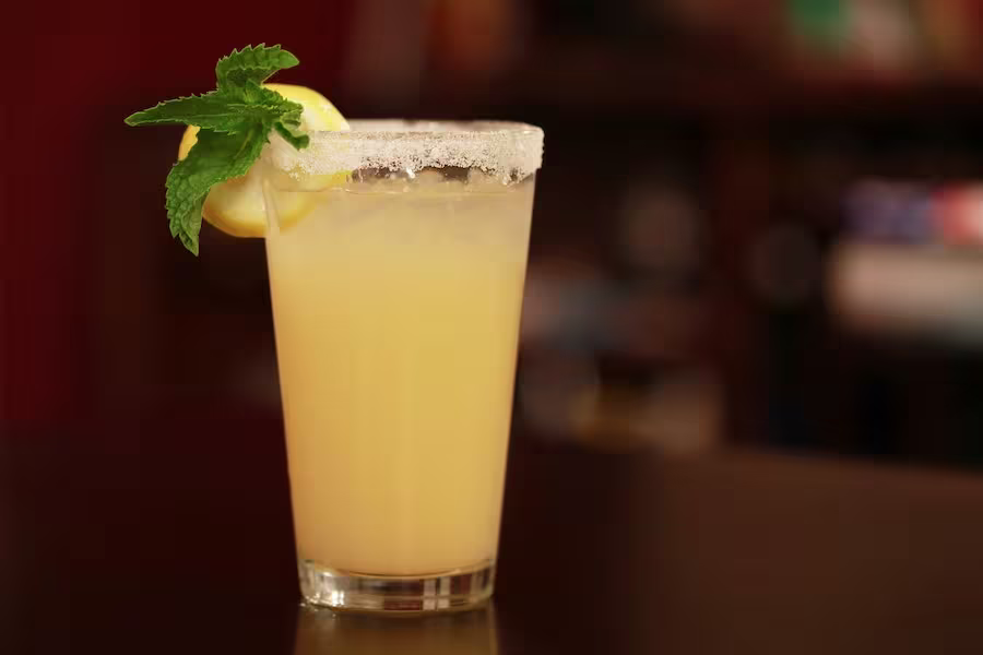 A glass of yellow lemonade with a sugared rim, garnished with a lemon slice and mint leaves, sits on a dark surface.
