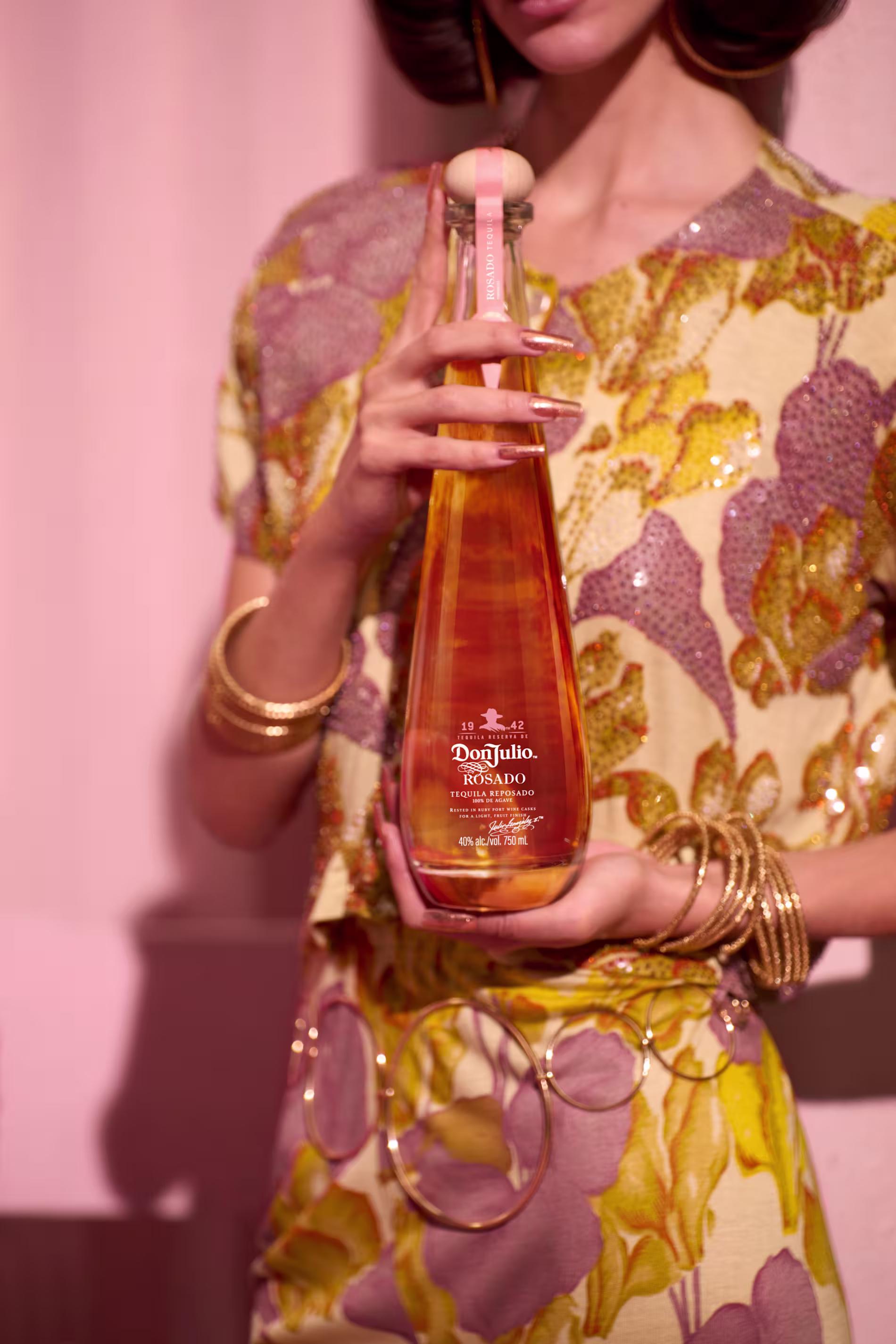 Woman in a floral dress holds a tall bottle of Don Julio Rosado tequila, with gold bracelets on her wrist and pink lighting in the background.