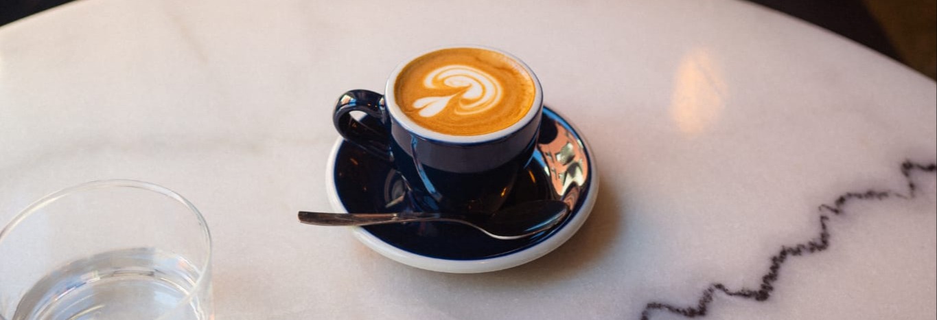 Café con arte latte en taza azul marino sobre mesa de mármol blanco veteado, acompañado de un vaso de agua y cucharilla en Trava de Noche.