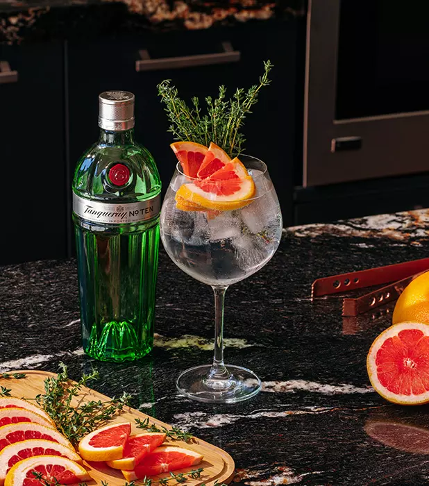 A green bottle of Tanqueray gin next to a gin and tonic garnished with grapefruit slices and thyme, on a dark marble counter with grapefruit and a wooden tray.