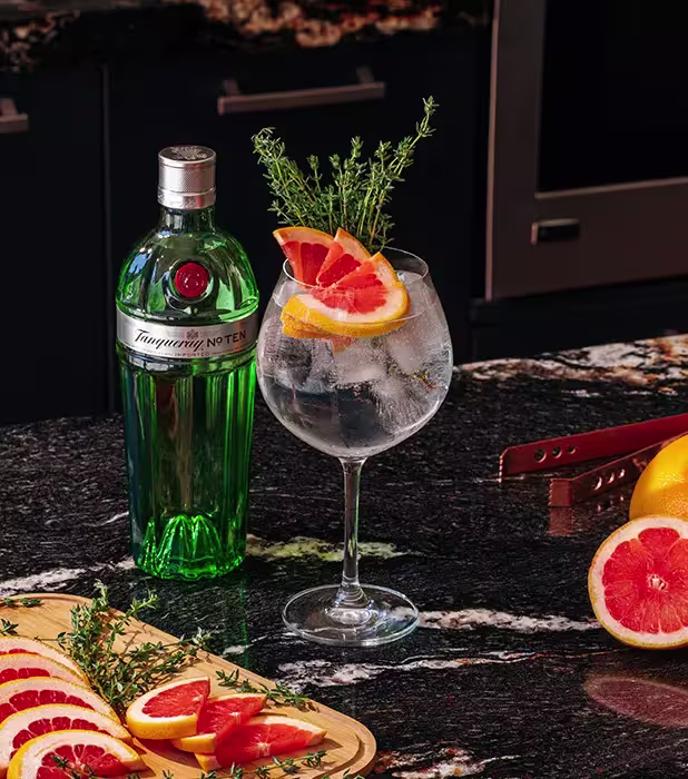 A green bottle of Tanqueray gin next to a gin and tonic garnished with grapefruit slices and thyme, on a dark marble counter with grapefruit and a wooden tray.