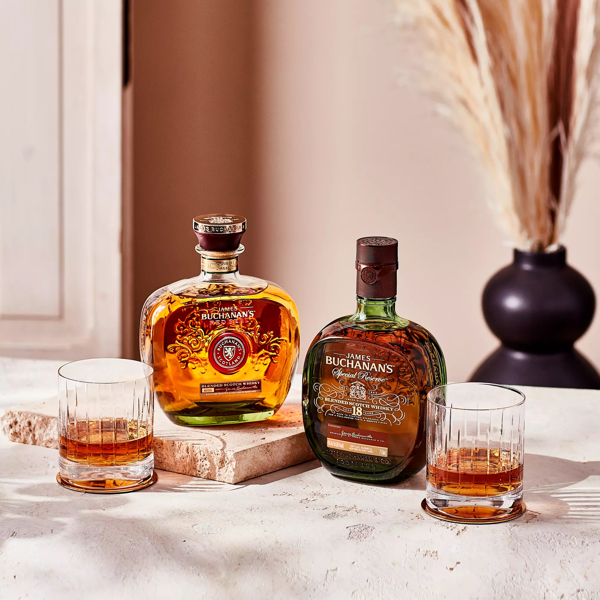 Two bottles of Buchanan’s whisky and two filled glasses are placed on a stone surface with a vase of dried grass in the background.