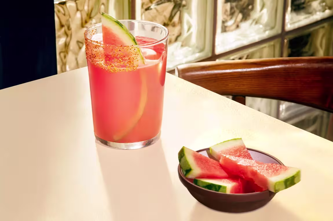 A glass of pink watermelon drink garnished with a watermelon slice and chili powder sits on a table next to a small bowl of watermelon wedges.