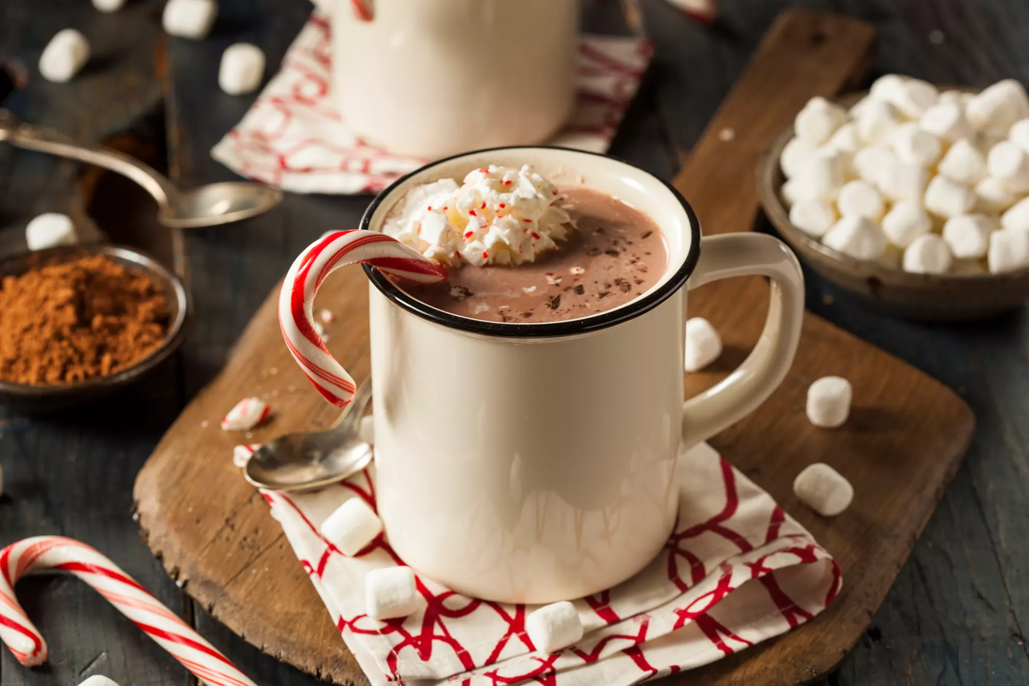 A mug of hot chocolate with whipped cream and a candy cane, surrounded by mini marshmallows, cocoa powder, and another mug in the background.