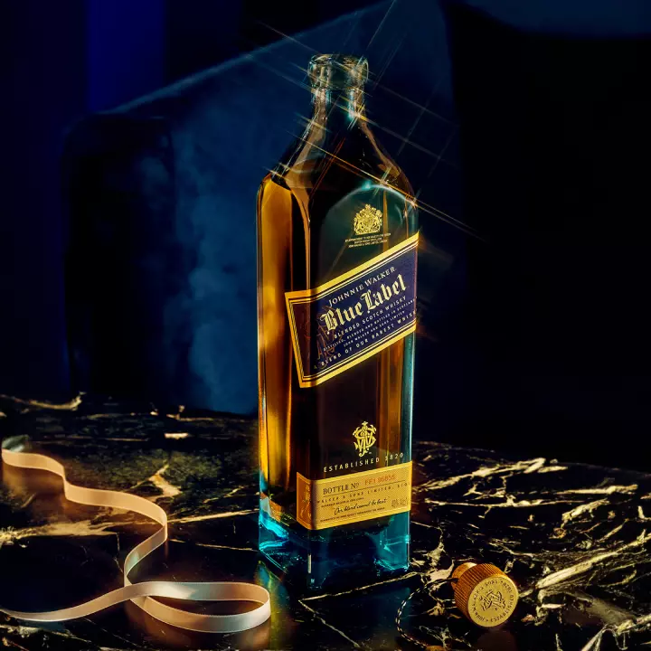 A bottle of Johnnie Walker Blue Label whisky stands on a marble surface next to its cap and a ribbon, with a dark, blurred background.