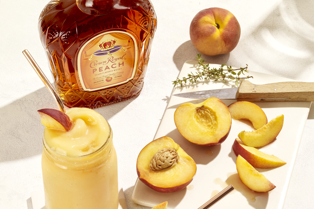 A bottle of Crown Royal Peach whiskey, a mason jar with a peach frozen drink, whole and sliced peaches, a knife, and a sprig of thyme on a white surface.