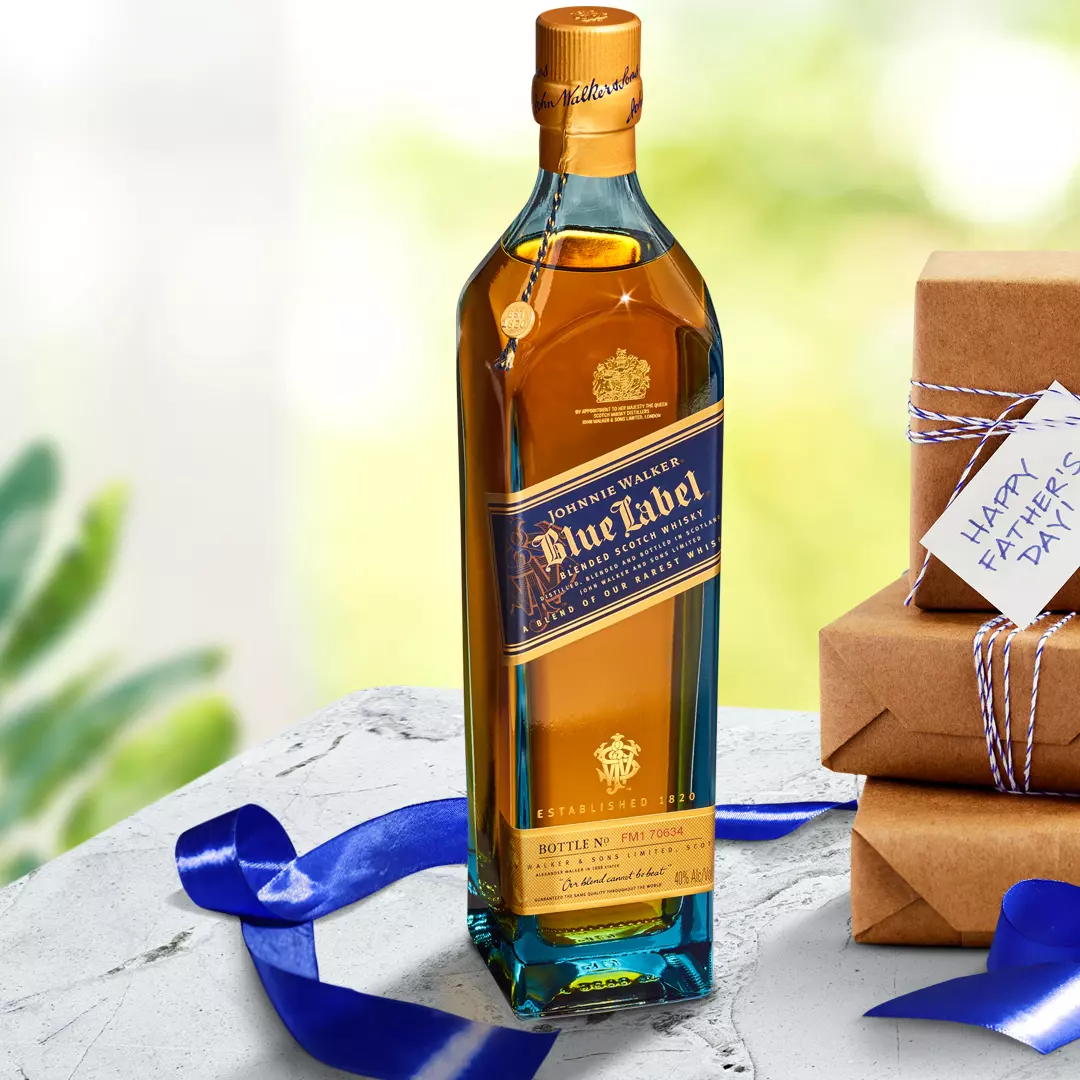A bottle of Johnnie Walker Blue Label whisky stands on a stone surface next to stacked gift boxes and a blue ribbon, with a Happy Fathers Day tag visible.