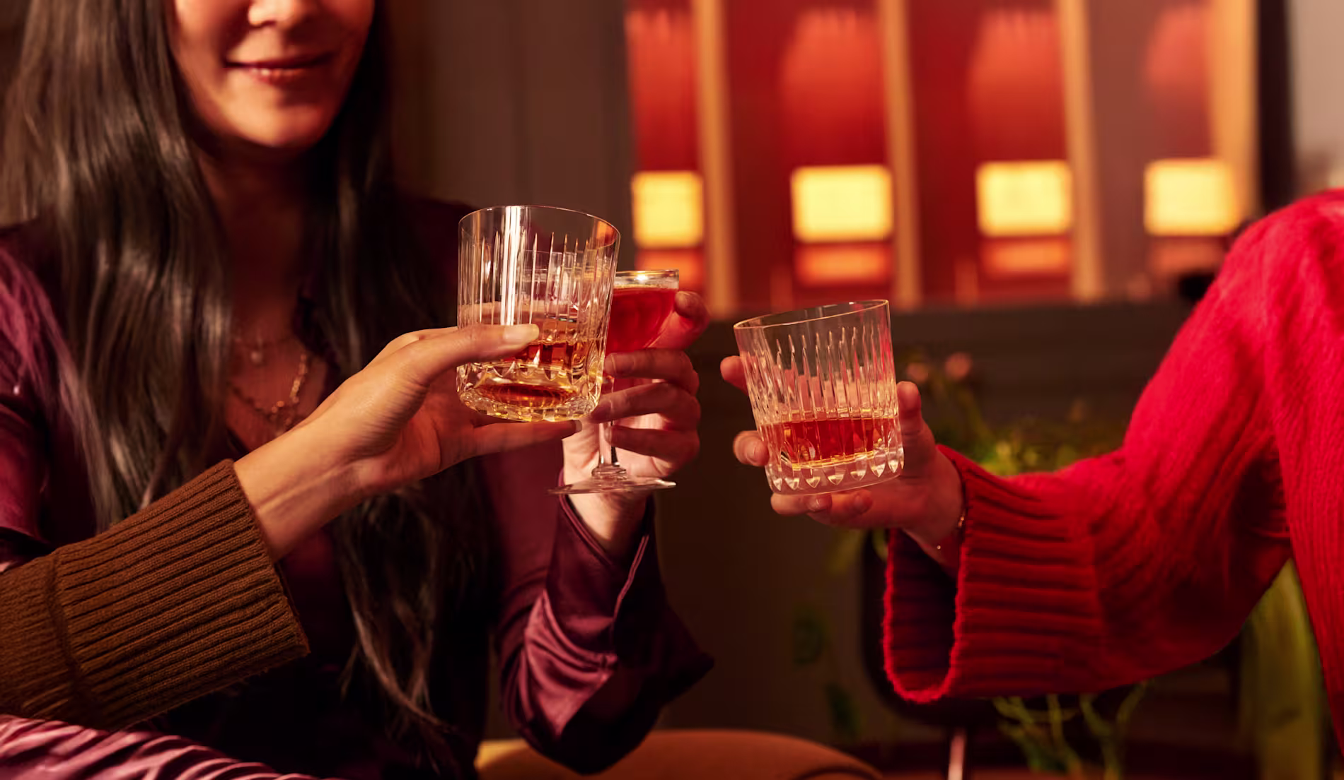 Three people clink glasses of whiskey and a cocktail in a warmly lit setting, with only their hands and parts of their upper bodies visible.