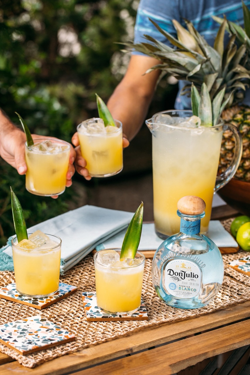 A pitcher and glasses of a yellow tropical cocktail with pineapple garnish are on a table with a bottle of Don Julio tequila; two people are serving drinks.