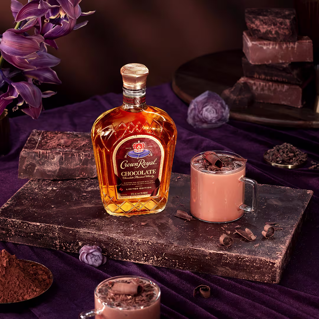 A bottle of Crown Royal Chocolate whiskey is placed on a stone slab with two mugs of hot chocolate, chocolate bars, cocoa powder, and purple flowers arranged nearby.