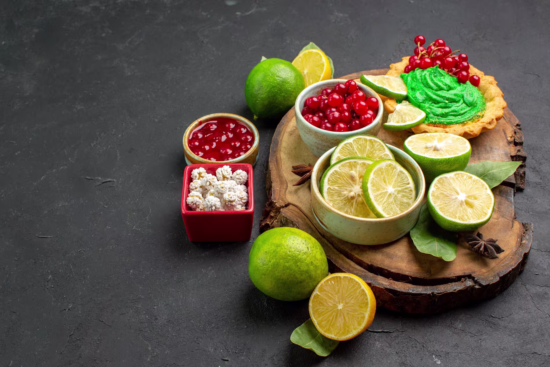 Assorted fresh limes, red currants, a green tart, and bowls of berries and candied treats are arranged on a wooden board and dark surface.