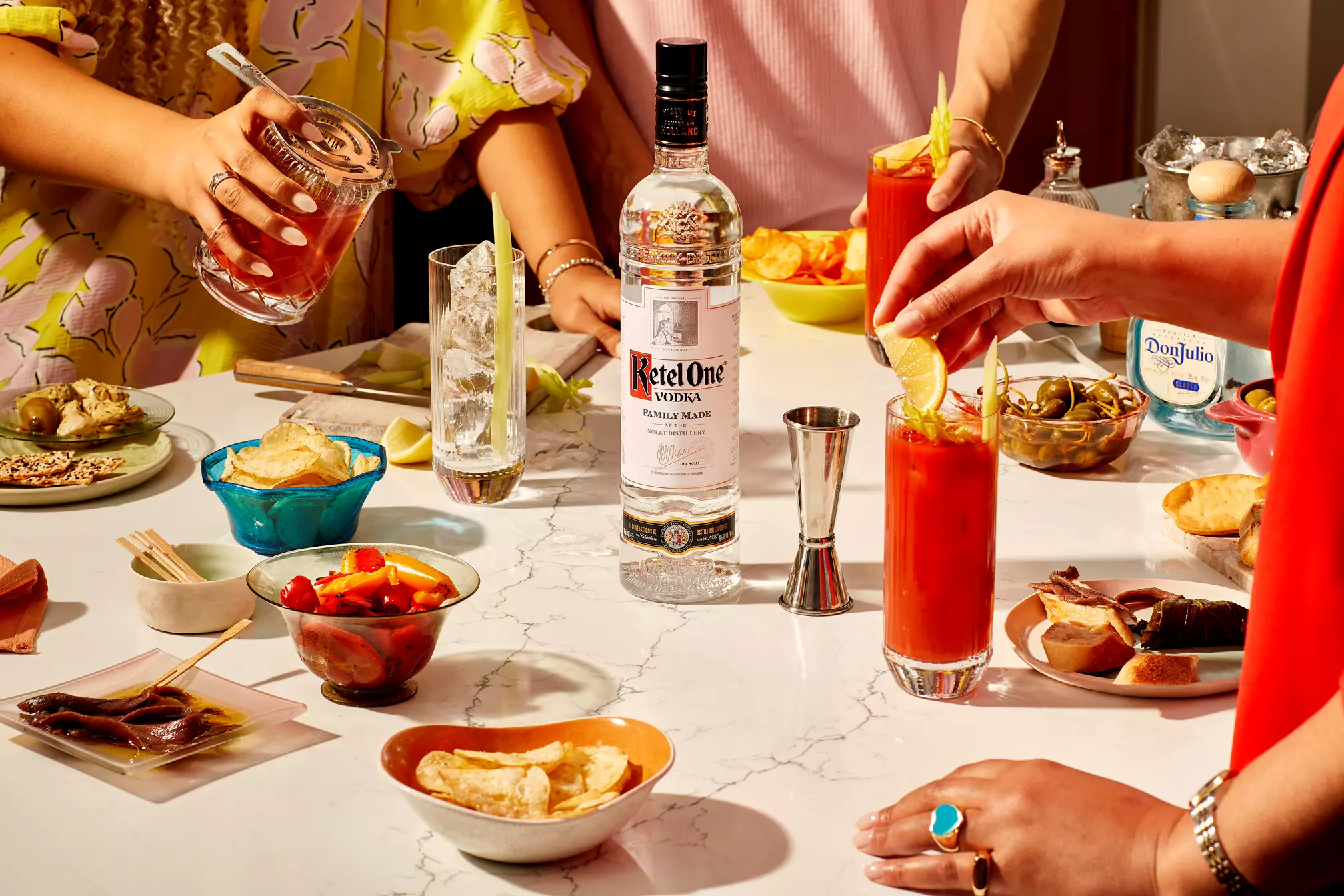 A group of people prepare and enjoy cocktails with a bottle of Ketel One vodka on a marble table surrounded by snacks and garnishes.