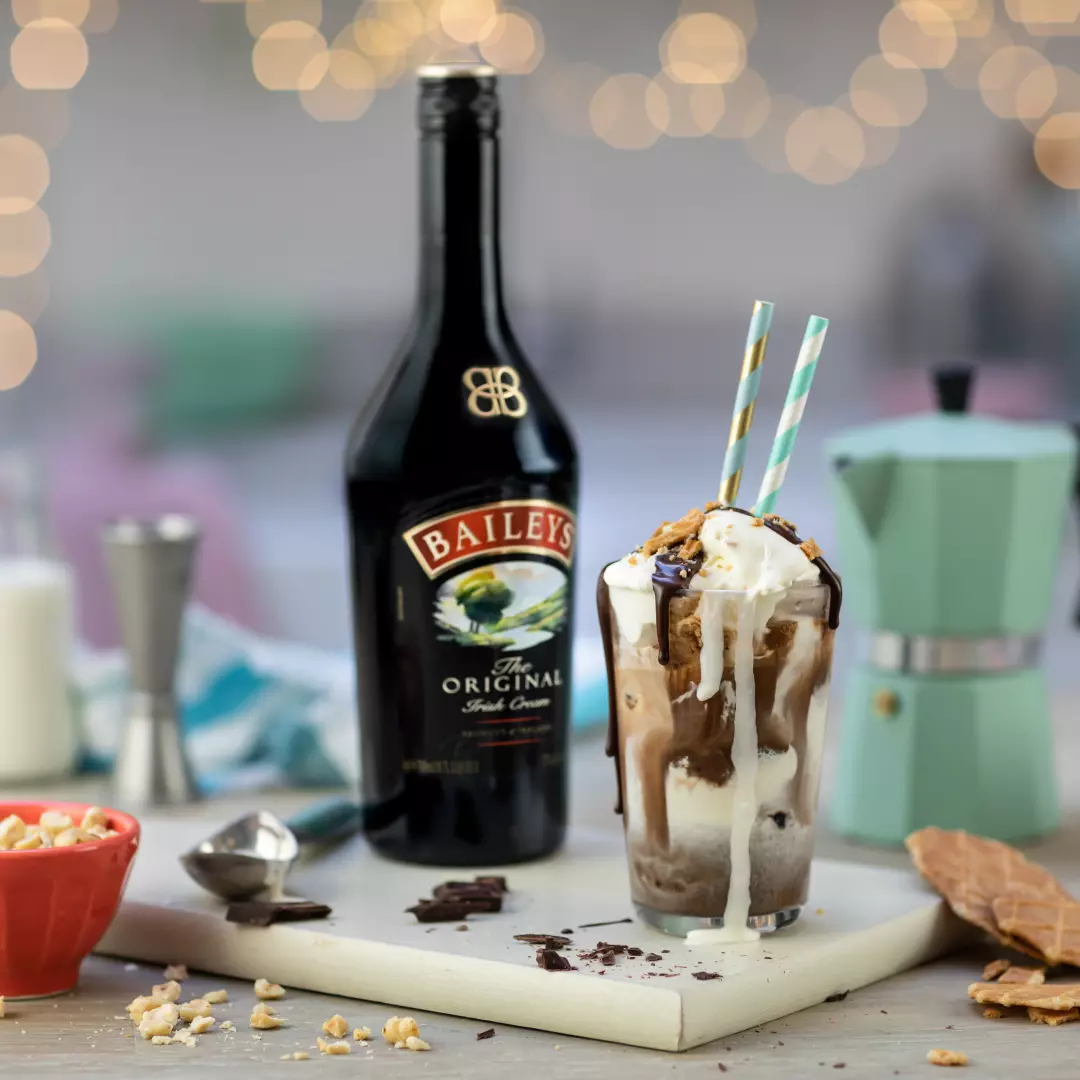 A bottle of Baileys Irish Cream stands behind a glass of ice cream float with straws, surrounded by toppings and a green moka pot on a white board.