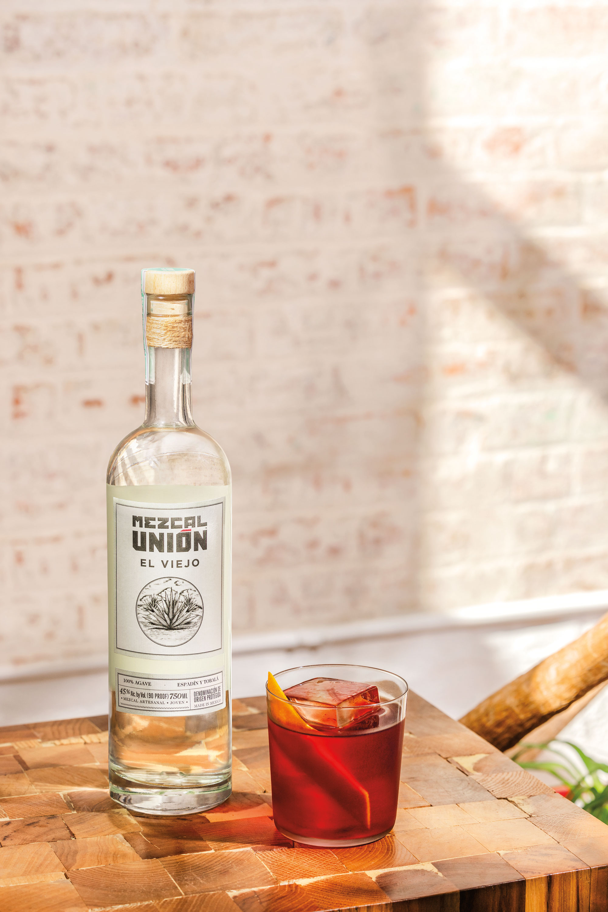 A bottle of Mezcal Unión El Viejo and a glass of red cocktail with orange garnish on a wooden table in sunlight.