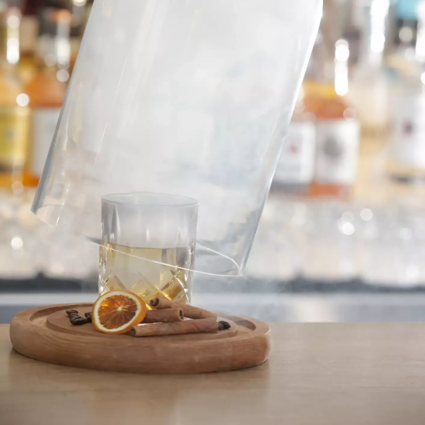 A hand lifts a glass dome filled with smoke from a cocktail on a wooden tray, garnished with a dried orange slice and spices. Bottles are blurred in the background.