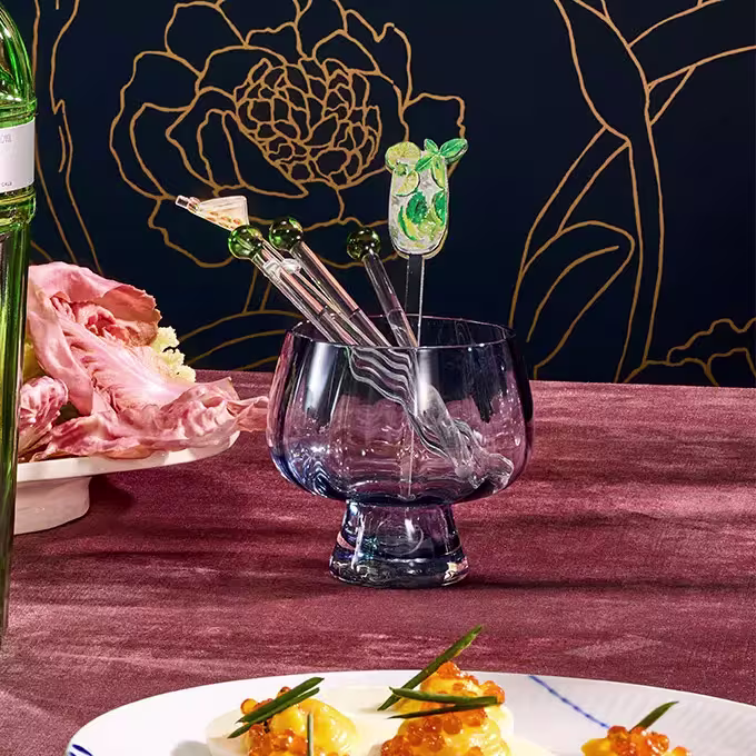 A blue glass goblet with three decorative cocktail stirrers sits on a pink tablecloth, with food and a green bottle nearby; floral wallpaper in the background.