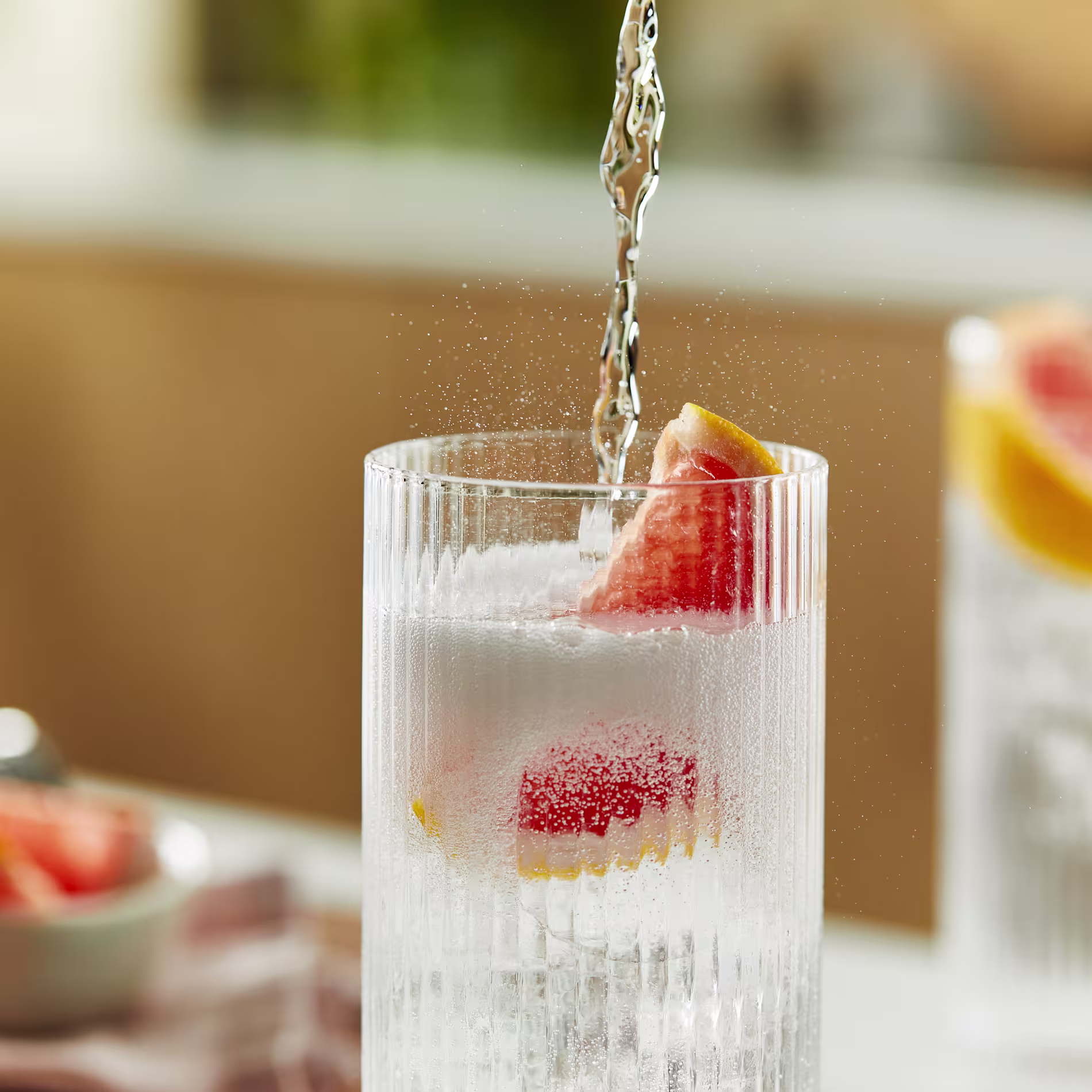A gin and tonic cocktail sits on a kitchen counter, with a pink grapefruit wedge sitting inside the highball glass. Tonic is poured from a height into the glass and it fizzes.