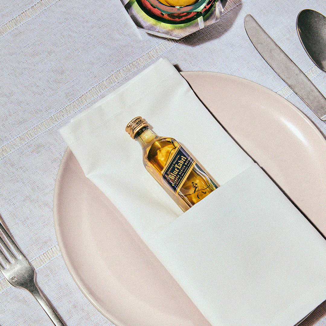 A small bottle of Ballantines whisky is placed in a folded napkin on a pink plate set with a fork, knife, and spoon on a white tablecloth.