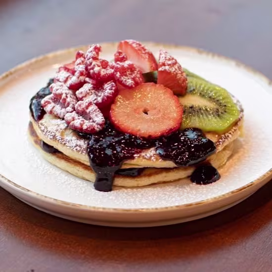 Torre de pancakes con sirope de frutos rojos, frambuesas con azúcar glas, fresas y rodajas de kiwi sobre un plato blanco con bordes dorados.