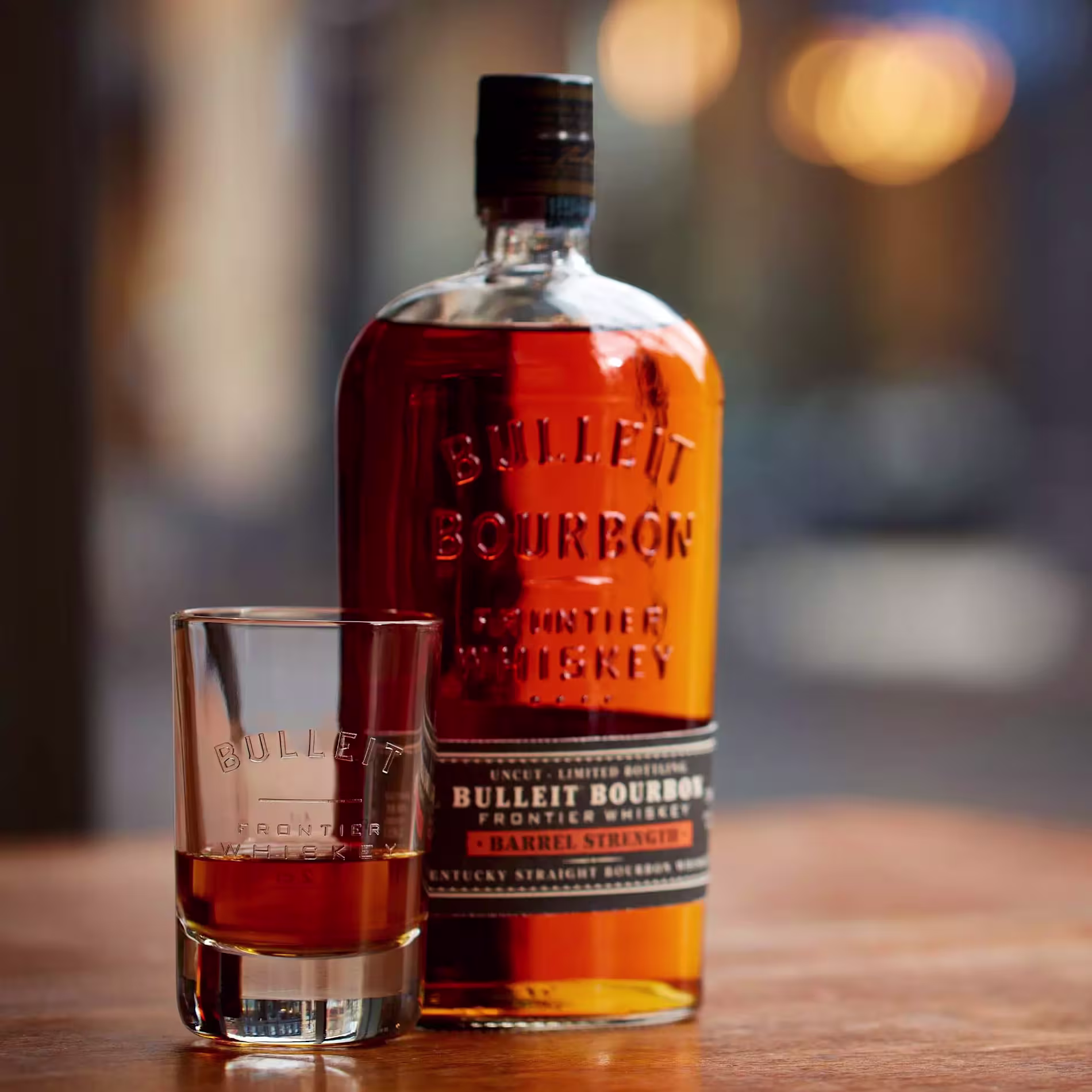 A bottle of Bulleit Bourbon Frontier Whiskey and a filled glass are placed on a wooden surface with a blurred background.