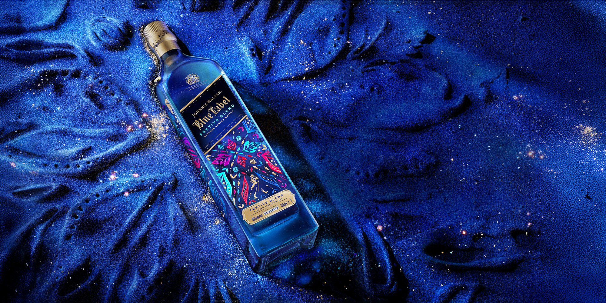Johnnie Walker Blue Label Festive Blend bottle displayed on a shimmering blue textured surface with glittering accents. The bottle features vibrant floral and festive artwork in pink, blue, and gold, illuminated by dramatic lighting for a premium holiday presentation.