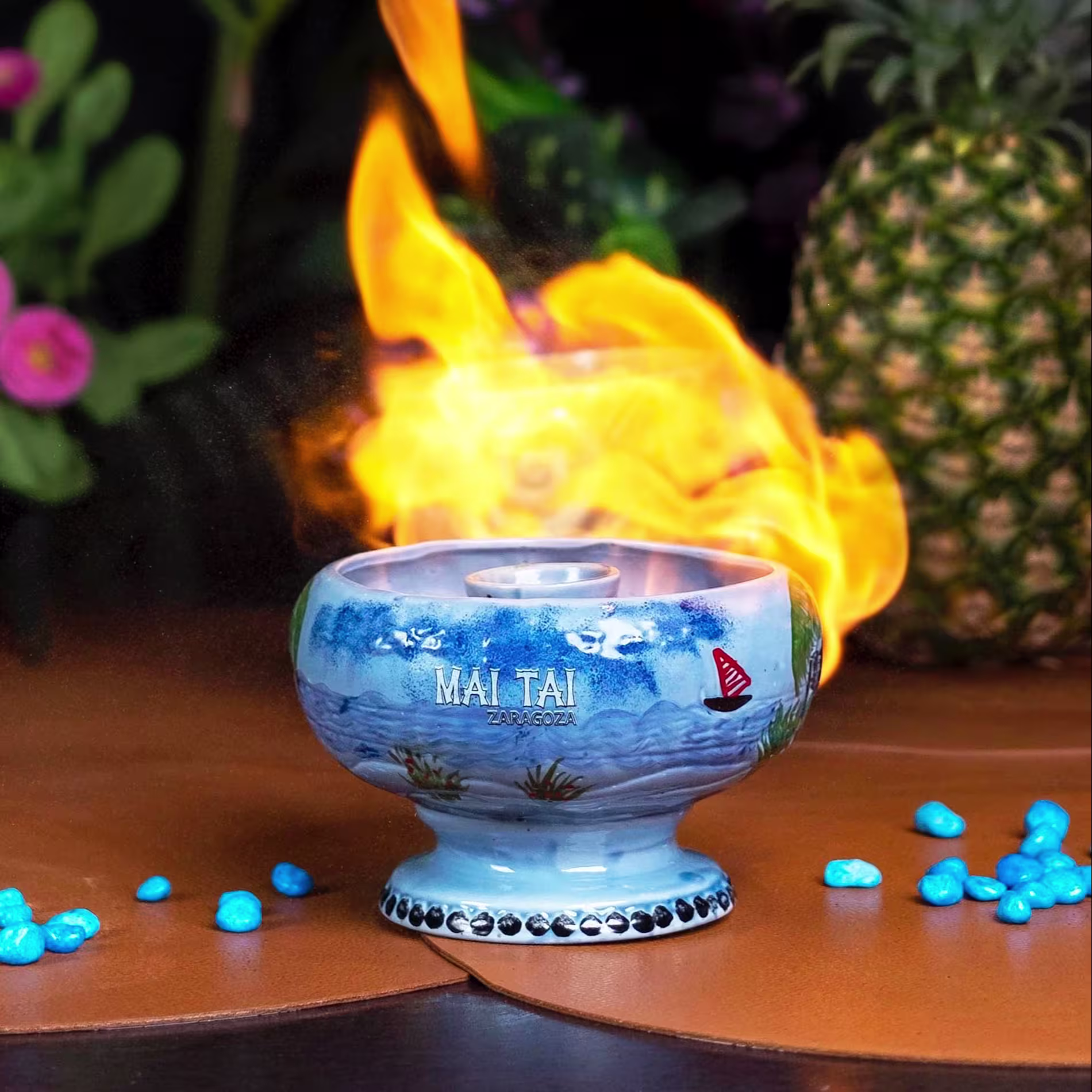 Cóctel servido en cuenco cerámico con centro en llamas, decorado con piedras azules y piña al fondo.