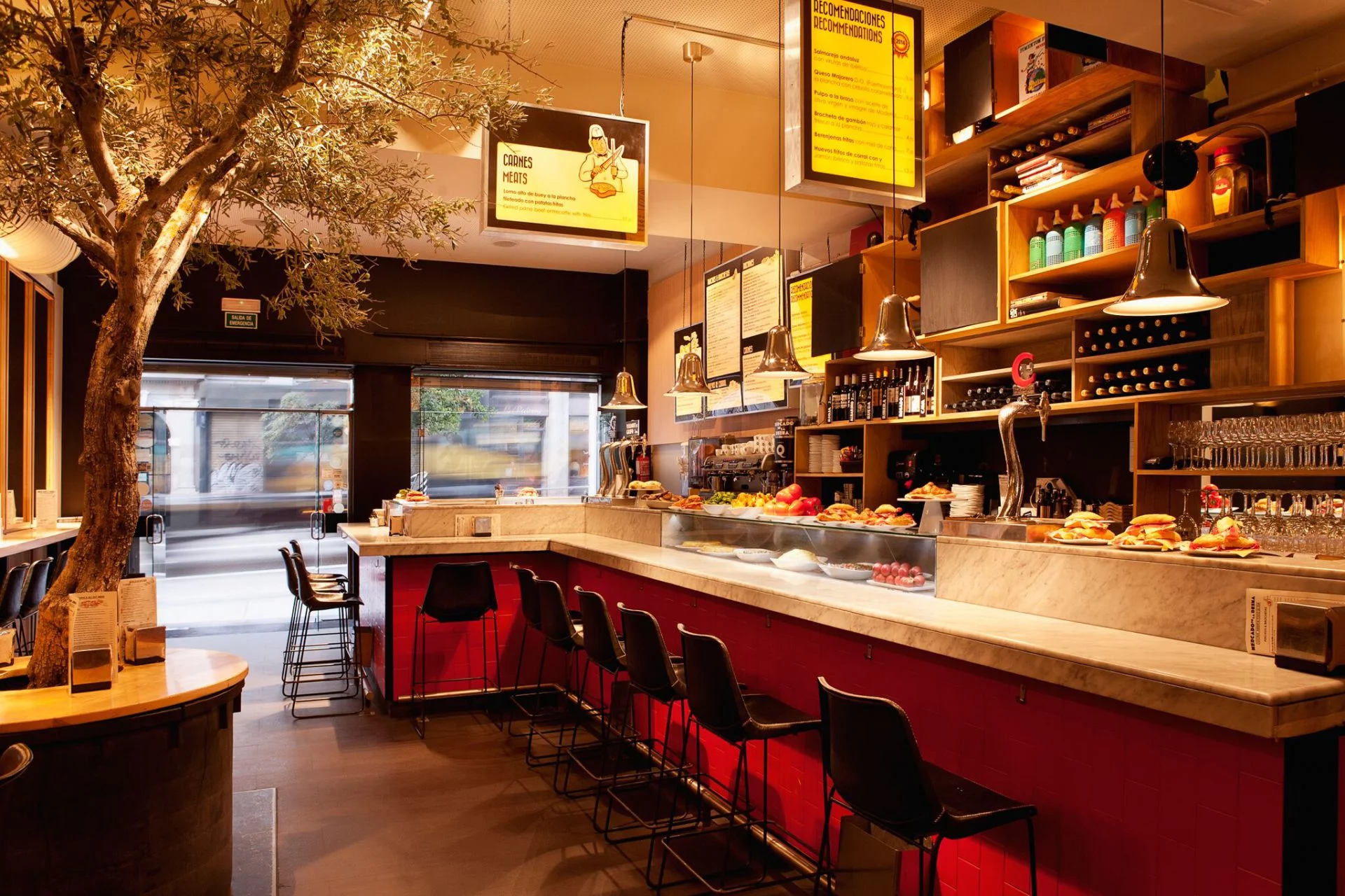 Barra de Diurno con un diseño moderno y acogedor. Resalta un árbol decorativo en el interior, acompañado de iluminación cálida y tapas apetitosas.