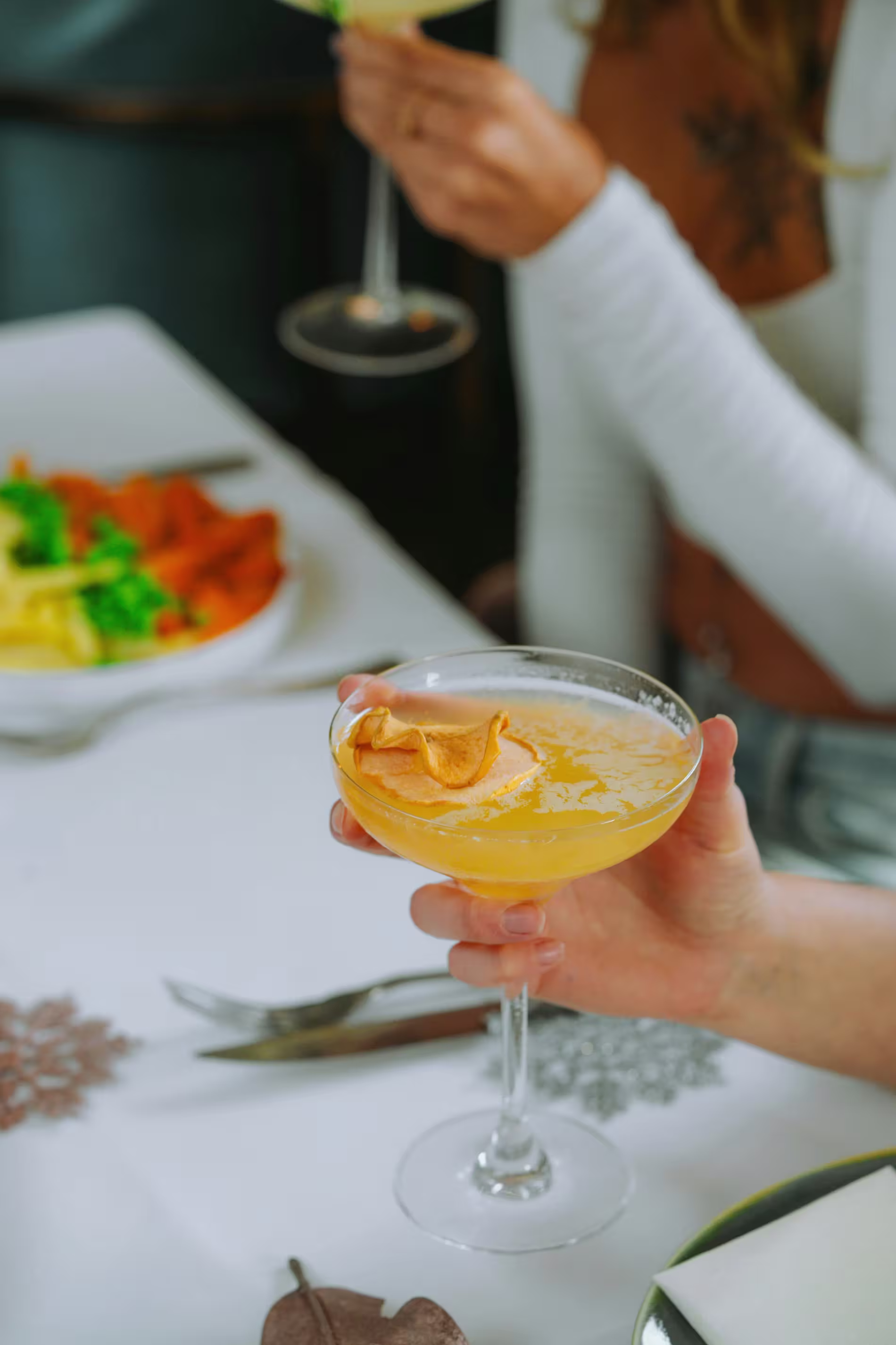 A person holds a cocktail garnished with a dried citrus slice at a table set with cutlery and a plate of food in the background.