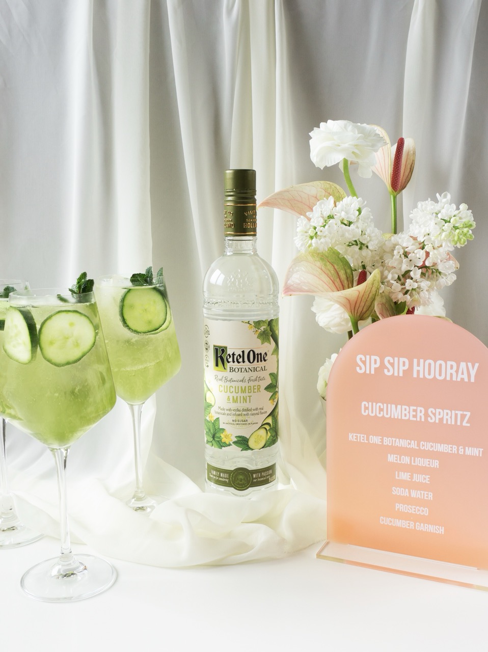 A bottle of Ketel One Botanical Cucumber & Mint stands beside three cucumber spritz cocktails, a pink menu card, and a flower arrangement on a white fabric background.