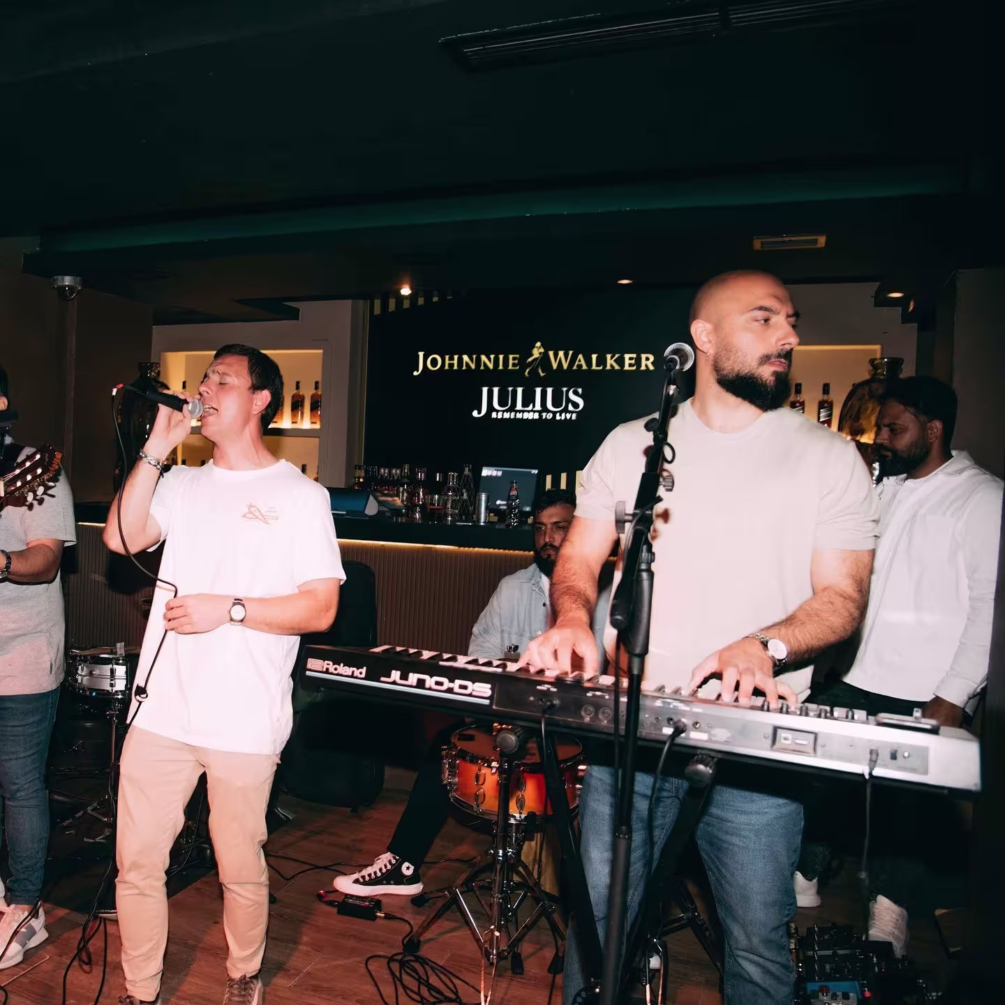 Concierto en vivo en Julius con banda tocando y cantante al micrófono. Ambiente animado y barra con visibilidad de Johnnie Walker.