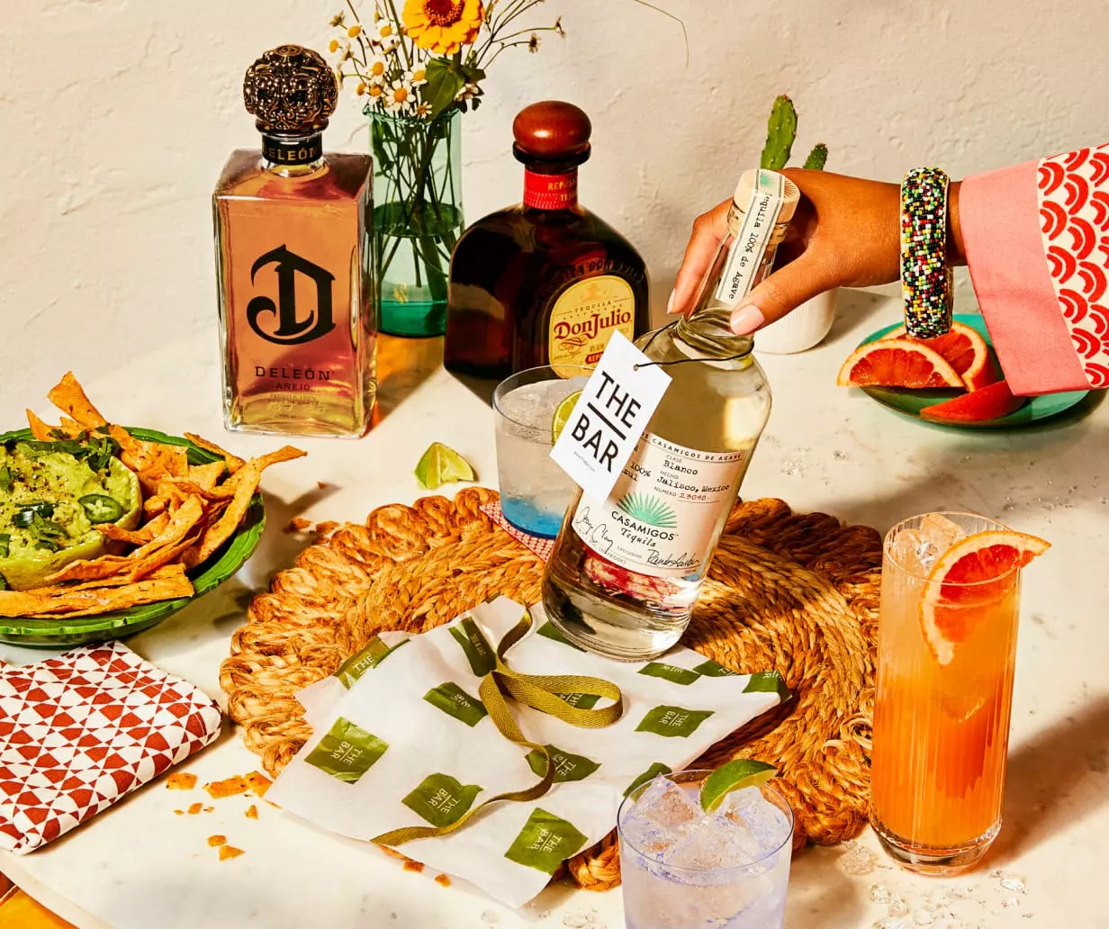 A hand holds a bottle of tequila over a woven placemat on a table set with drinks, chips, salsa, and two other tequila bottles. Decor and flowers are also visible.