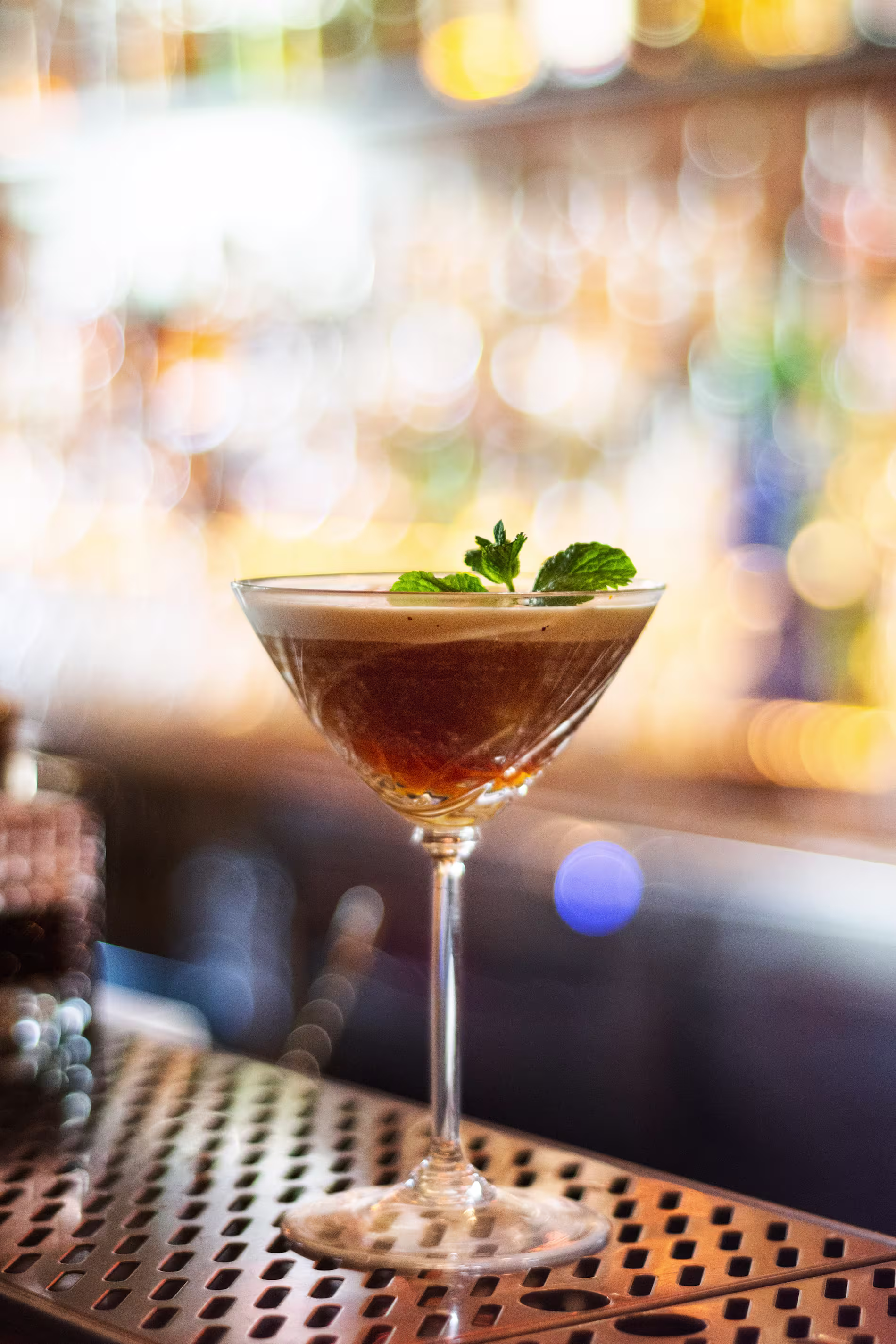 La imagen muestra un primer plano de un cóctel en una copa de martini, adornado con una ramita de menta en la parte superior. El cóctel está colocado en un mostrador de bar.