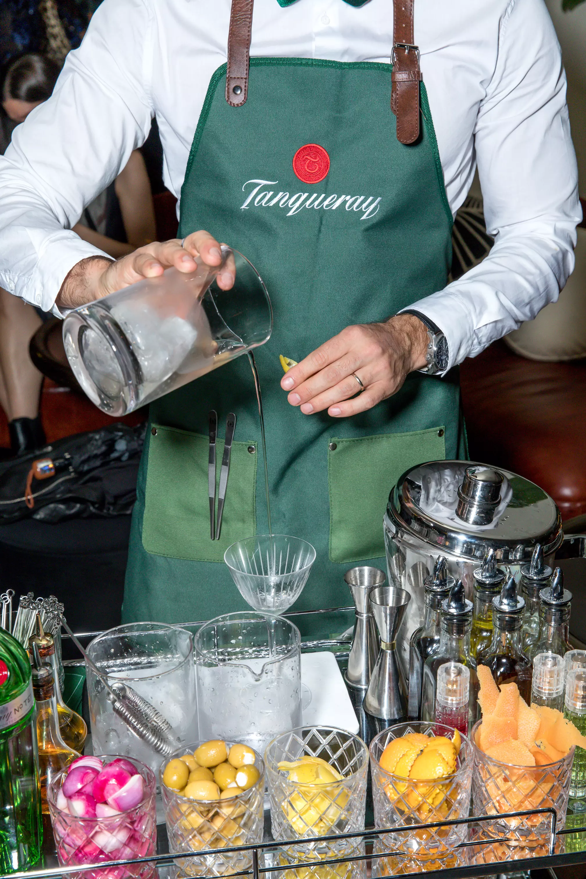 A bartender in a green Tanqueray apron pours a drink through a strainer into a cocktail glass, surrounded by bar tools, garnishes, and bottles.