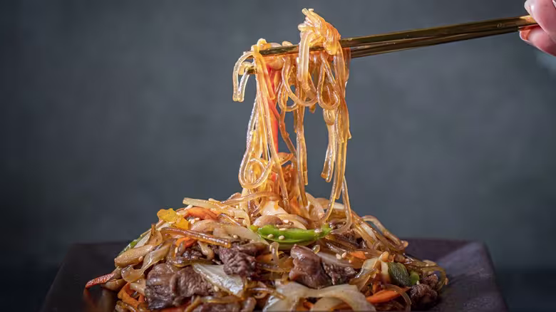 A hand uses chopsticks to lift glass noodles, vegetables, and meat from a plate of stir-fried noodle dish.