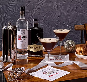 Two espresso martinis in cocktail glasses on a wooden table with bottles of Ketel One vodka and Mr Black coffee liqueur, napkins, a coffee press, a pastry, and decorative items.